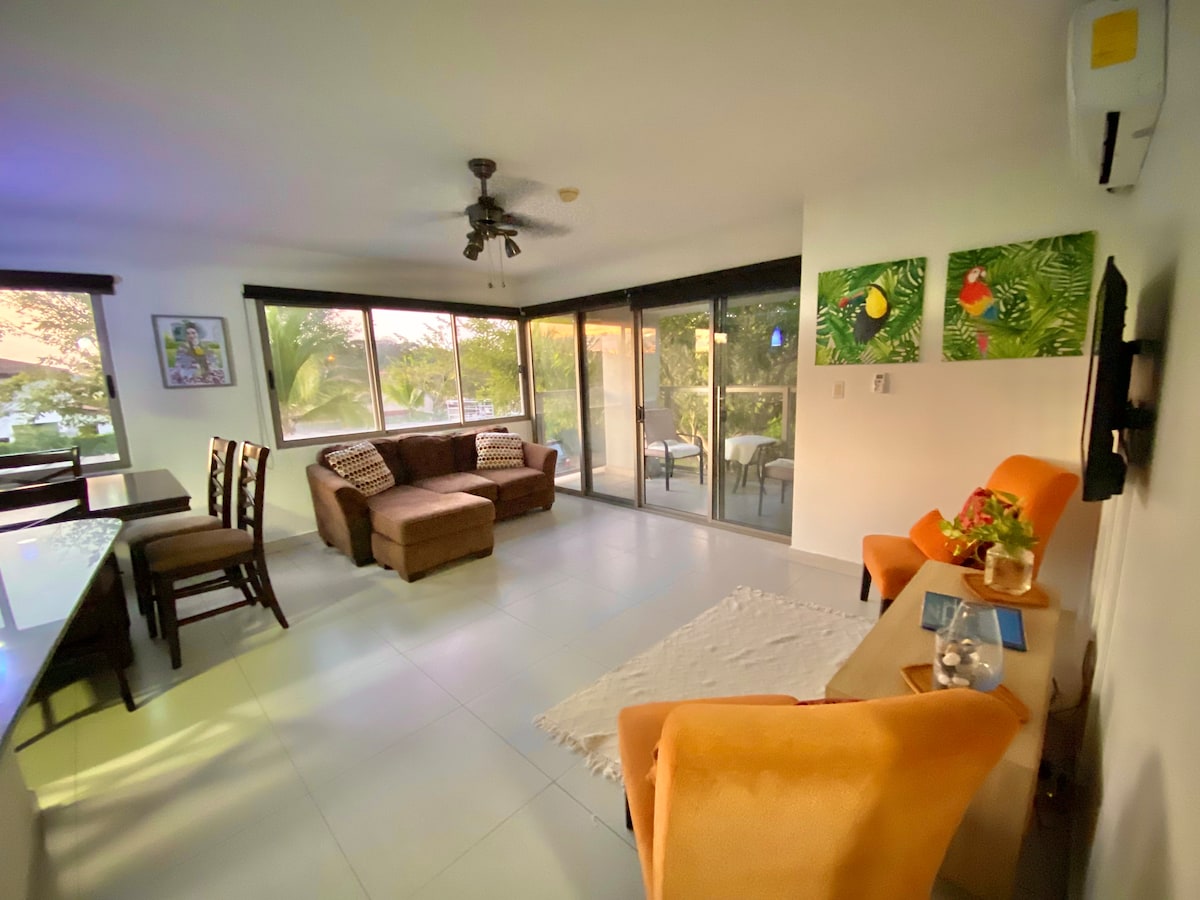 The living area features a blend of seating options, including a spacious sofa and bright orange armchairs. Large glass doors provide ample natural light and a view of the surrounding greenery. Artwork adorns the walls, adding a touch of color to the neutral palette.
