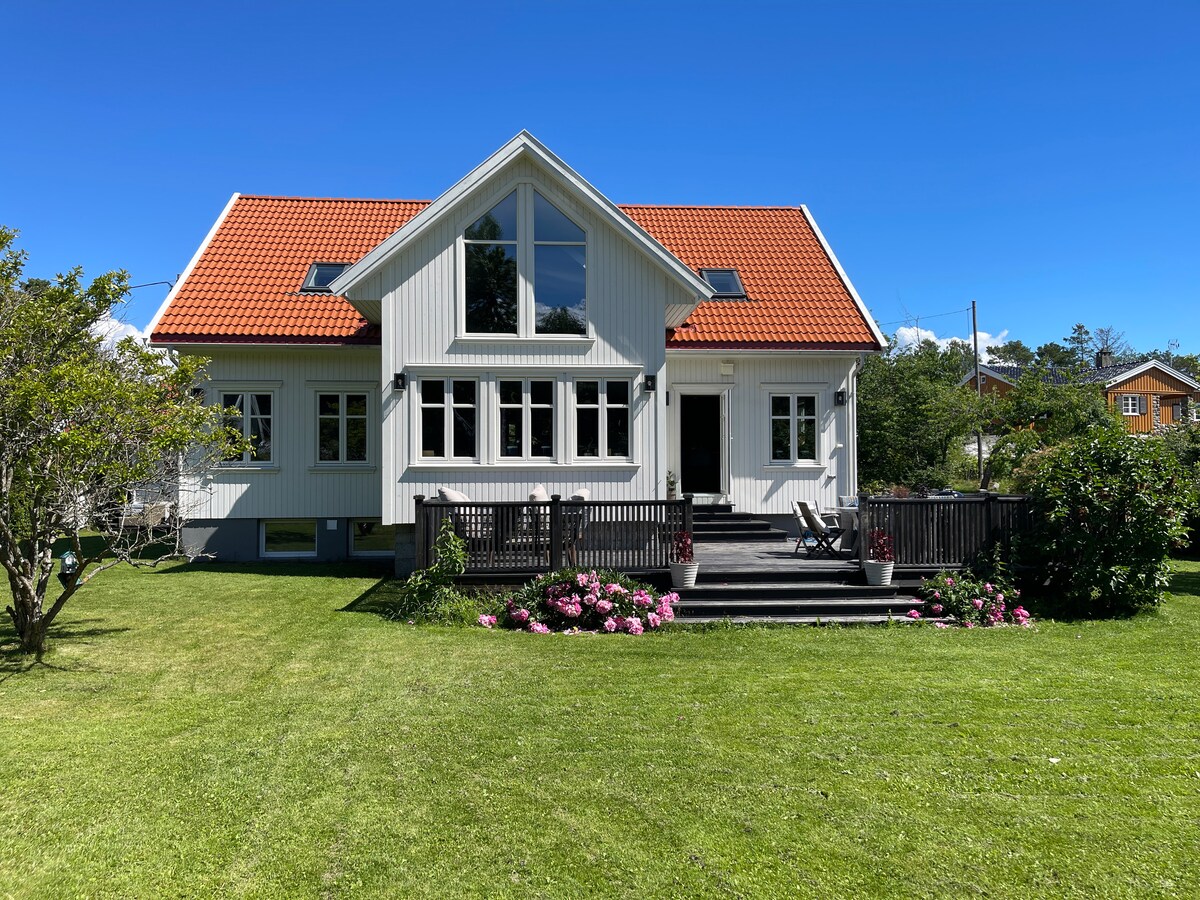 A two-story house features a light exterior with a red-tiled roof and large windows. A spacious deck is surrounded by lush green grass and flower beds, inviting outdoor relaxation. The setting is peaceful with a clear blue sky in the background.