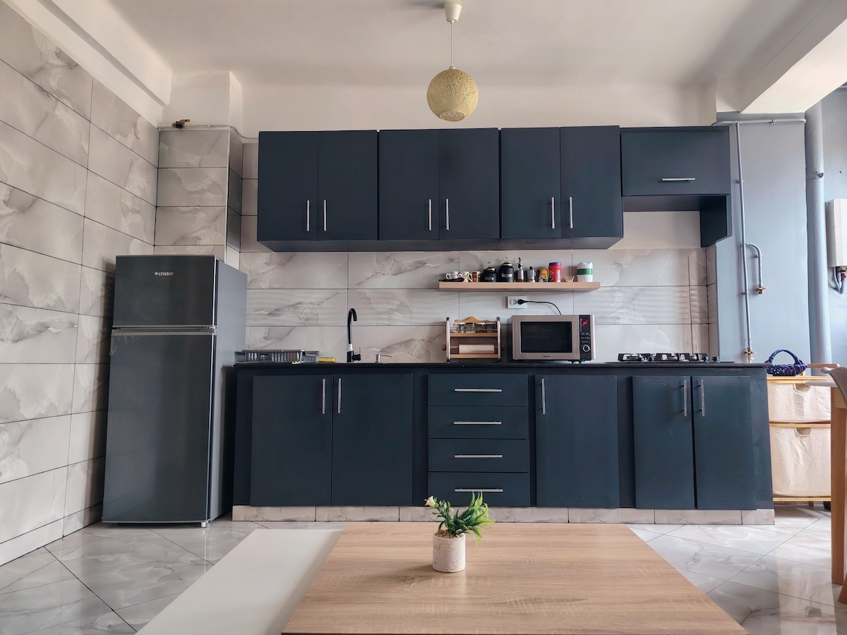 A modern kitchen is displayed, featuring dark cabinetry and a light marble backsplash. A refrigerator, microwave, and stove are integrated into the design. A wooden table is placed in the foreground, accompanied by a small potted plant, adding a touch of greenery to the space.