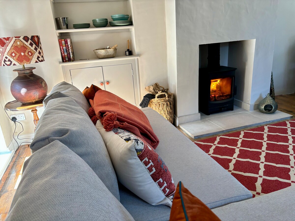 A cozy corner of the living area features a grey sofa adorned with decorative pillows and a warm throw. A wood-burning stove provides a gentle glow, while a patterned rug complements the space. Shelving displays various items, enhancing the inviting atmosphere.