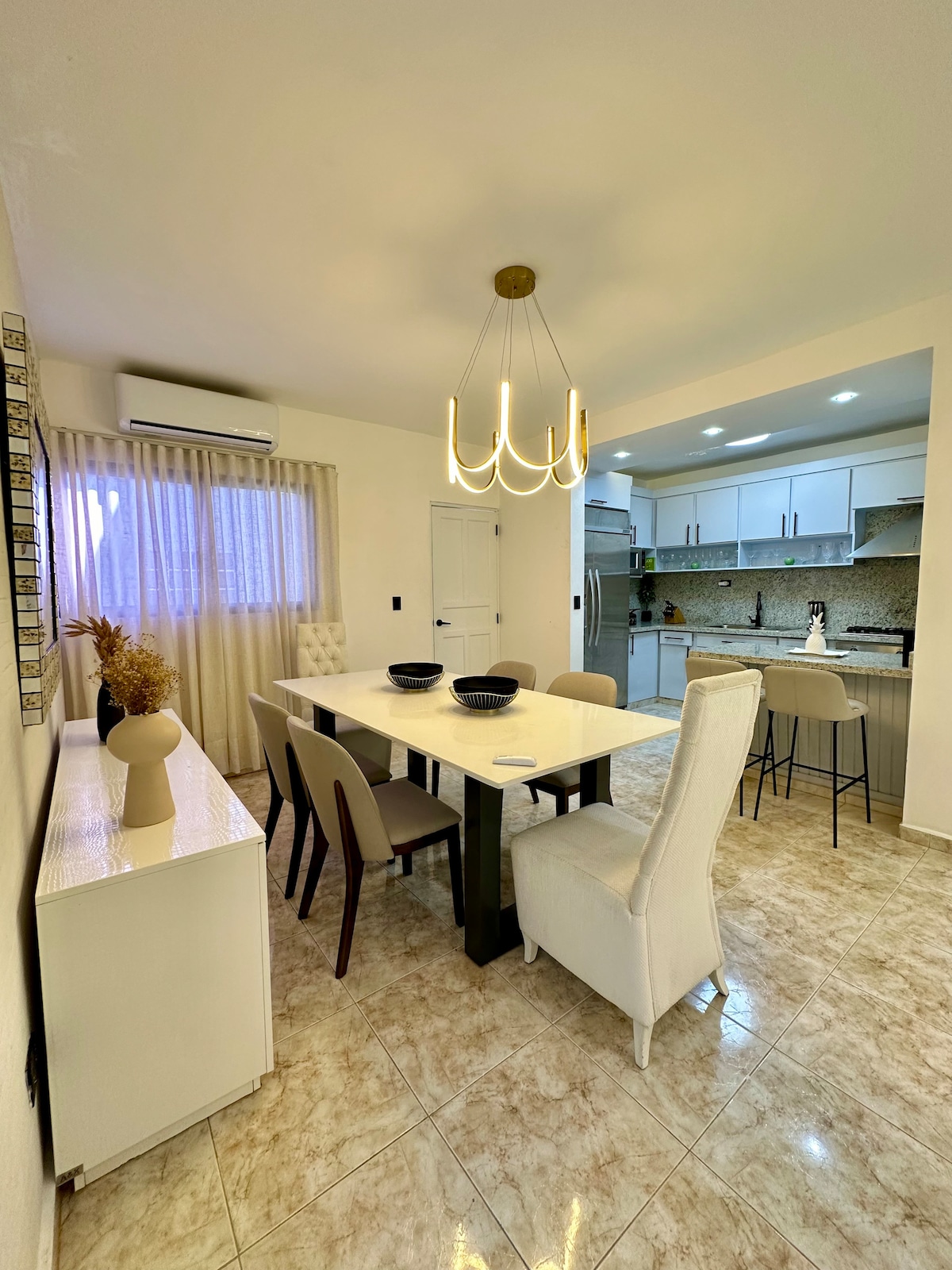 A dining area features a long table surrounded by mix of upholstered chairs. A modern light fixture hangs overhead, illuminating the space. Natural light enters through a window dressed with sheer curtains, highlighting the neutral-toned walls and elegant decor.
