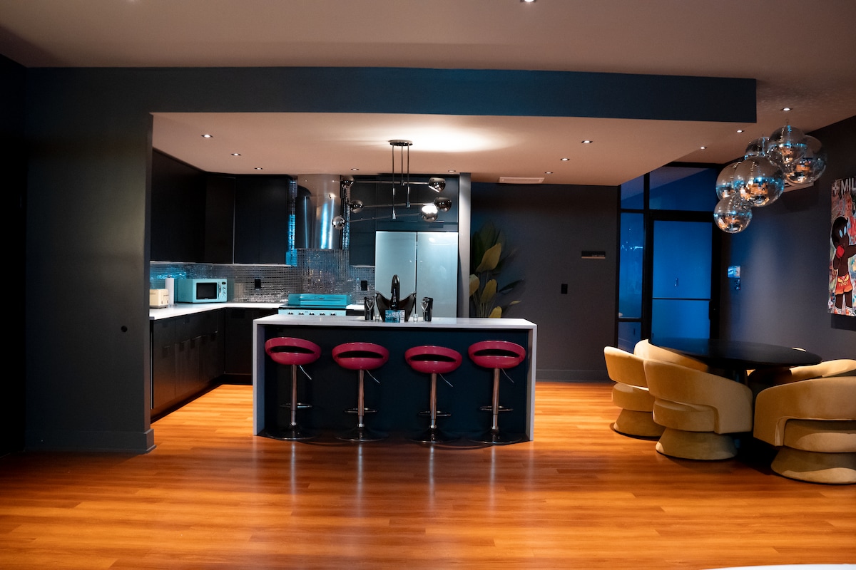 The modern kitchen features dark cabinetry and a spacious countertop, complemented by several sleek bar stools. A round dining table is situated nearby, adorned with comfortable seating. Lighting fixtures hang overhead, enhancing the contemporary design, while large windows provide natural light.