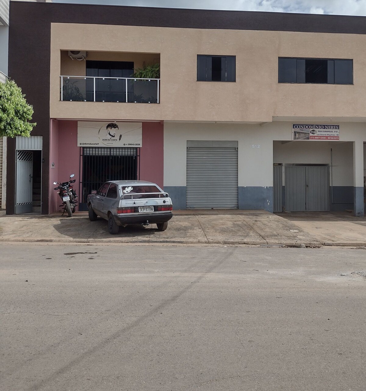 A multi-story building features a neutral-colored facade with windows on the upper level. A parked vehicle is visible in front, alongside a motorcycle. An entrance with signage is located on the ground floor, providing access to the space. The surrounding street is paved.