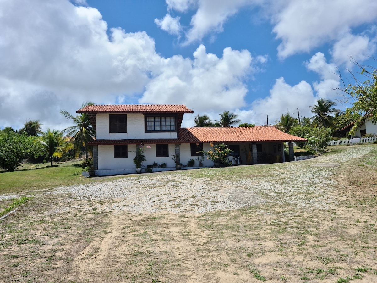 A two-story house with a tiled roof is surrounded by greenery. The exterior features a combination of white walls and dark window frames, providing contrast against the blue sky and fluffy clouds. A gravel path leads to the entrance, enhancing accessibility.