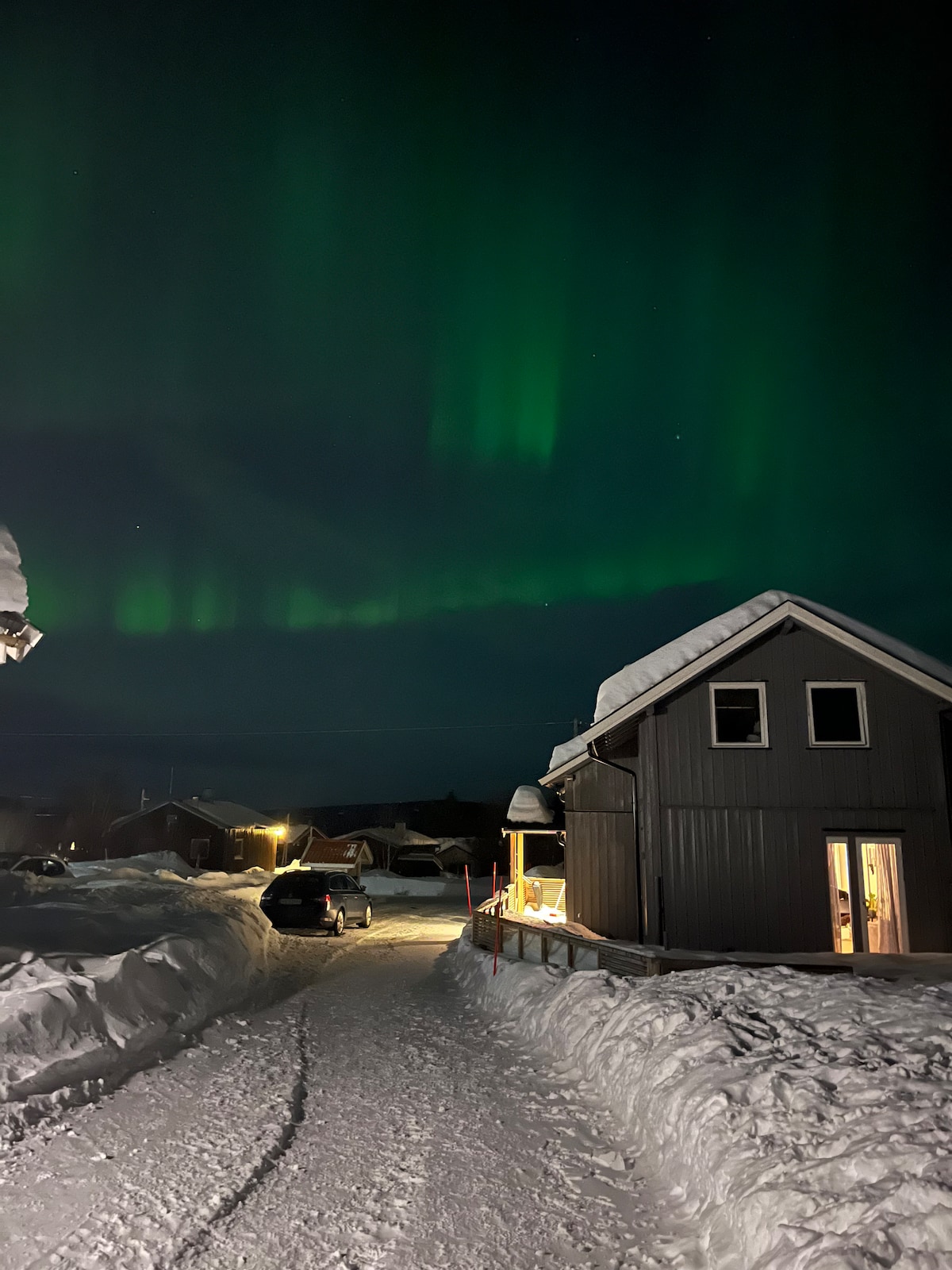 A charming house is situated along a snow-covered pathway, with vibrant green northern lights illuminating the night sky above. The structure features a well-maintained exterior and warm light emanates from the windows, creating a welcoming ambiance amidst the snowy landscape.