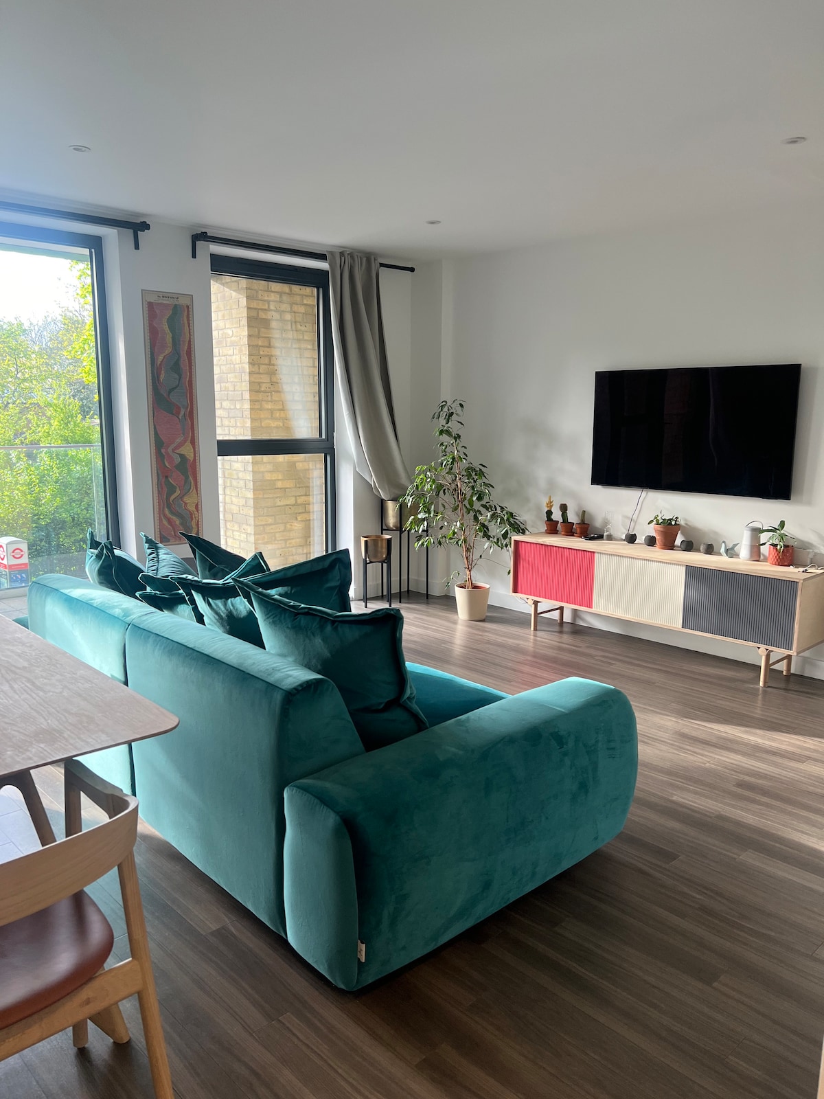 A spacious living area is furnished with a large teal sofa and a wooden dining table. Natural light enters through floor-to-ceiling windows, with greenery visible outside. A modern entertainment unit displays decorative items and a flat-screen television is mounted on the wall.