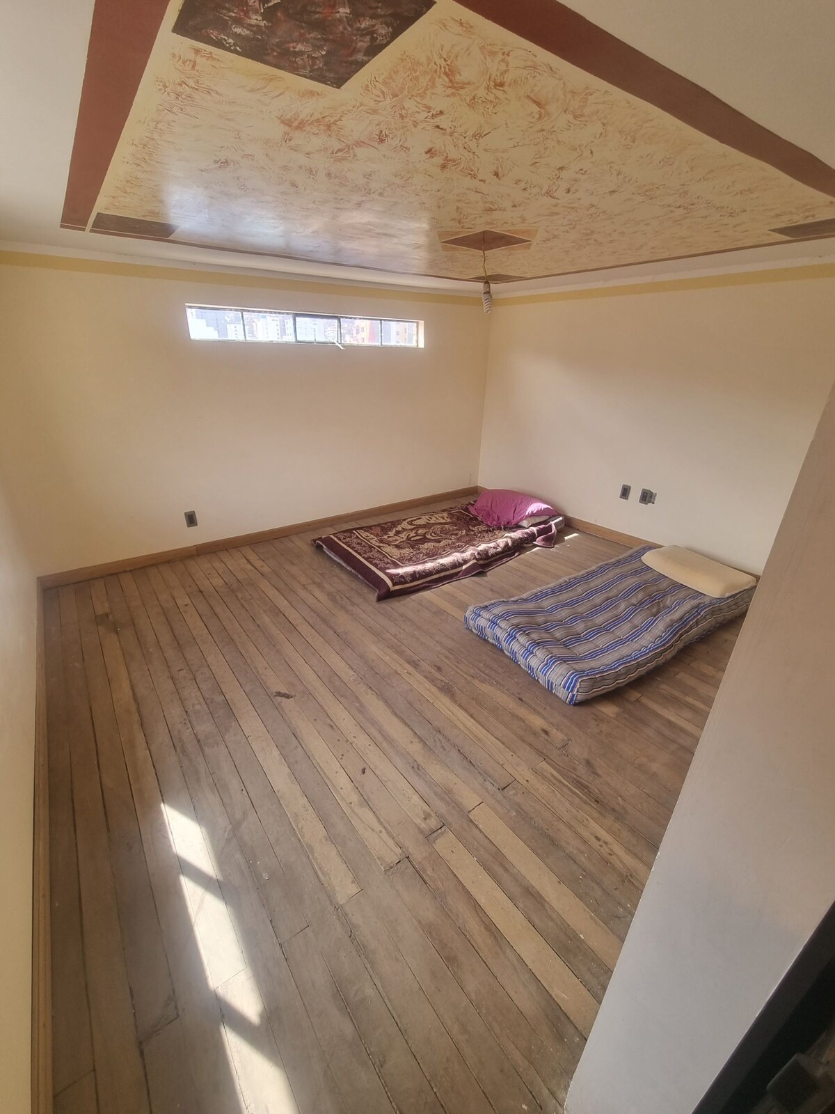 An expansive room features hardwood floors and a decorated ceiling. Two sleeping areas are present, each with a mattress on the floor: one is adorned with a patterned blanket, and the other is covered by a striped fabric. Natural light enters through multiple small windows.