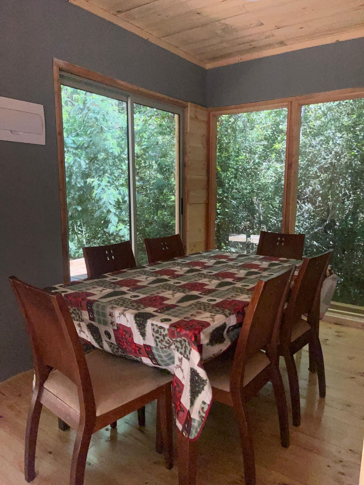 A dining area is presented with a large table covered by a patterned tablecloth. Surrounding the table are six wooden chairs. Large windows provide views of lush greenery, enhancing the natural light in the space.