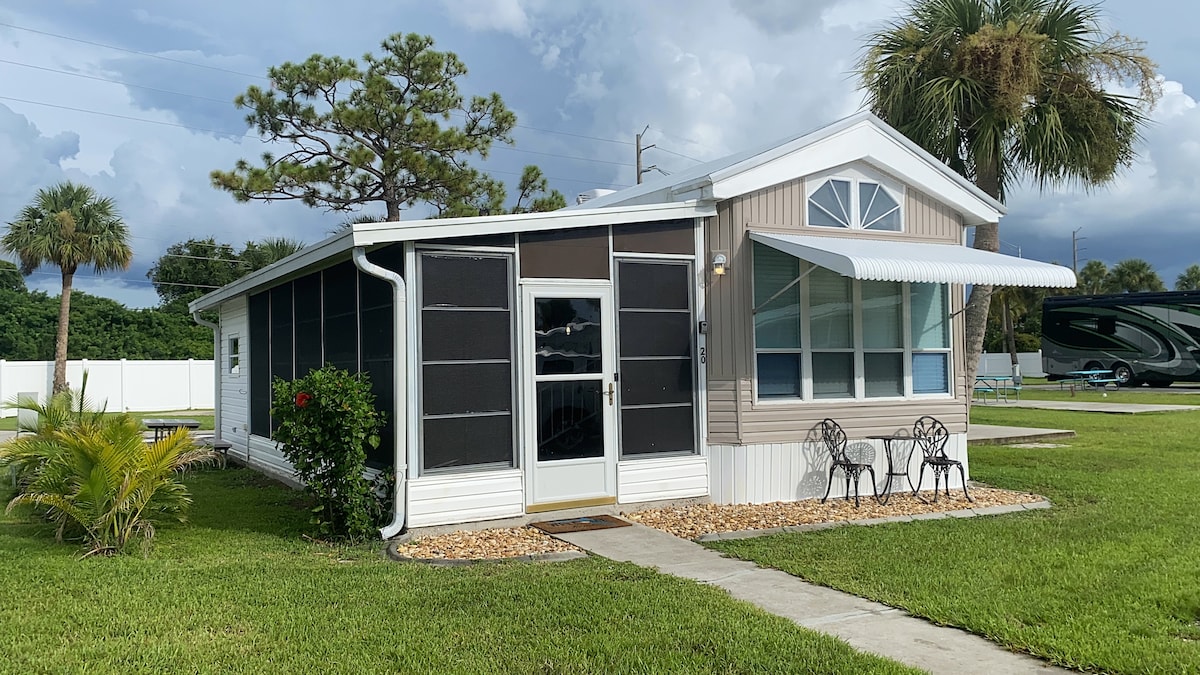 A newly remodeled tiny home presents a charming exterior, featuring a covered porch with large windows. Two chairs are placed outside under an awning, surrounded by manicured grass and tropical landscaping, including palm trees. The property is bordered by a decorative gravel walkway.