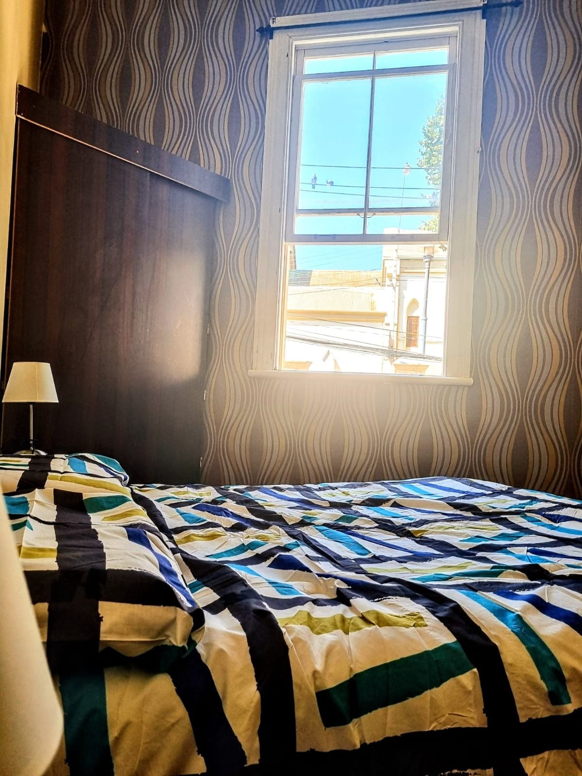 A bedroom is featured with a striped bedspread covering a spacious bed. A tall window allows natural light to filter in, providing views of the surrounding area. A small lamp is placed on a bedside table, complementing the patterned wallpaper.
