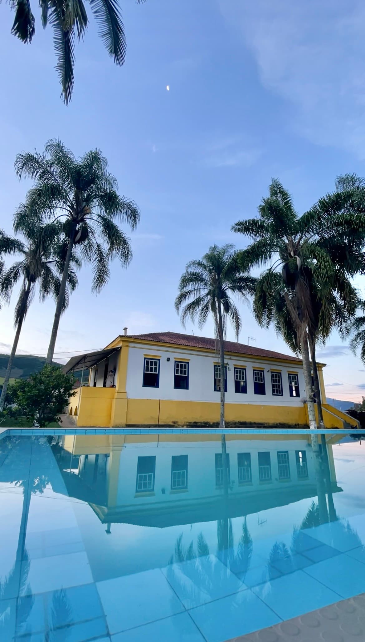 A charming building featuring a yellow exterior is reflected in a clear swimming pool. Lush palm trees surround the area, with a serene sky in the background, creating a tranquil setting. The structure includes multiple windows, allowing ample natural light to enhance its welcoming appearance.