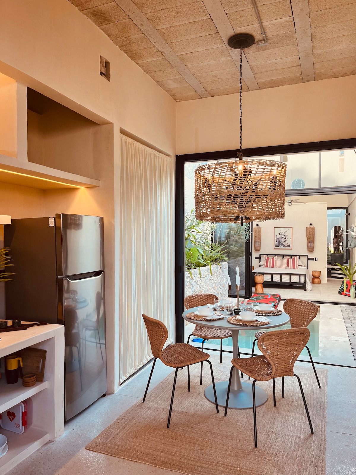 A modern dining area is featured, with a round table set for four. Stylish woven chairs surround the table, which is adorned with dishes and glassware. Soft lighting is provided by a large woven pendant lamp, complementing the sleek kitchenette visible in the background.
