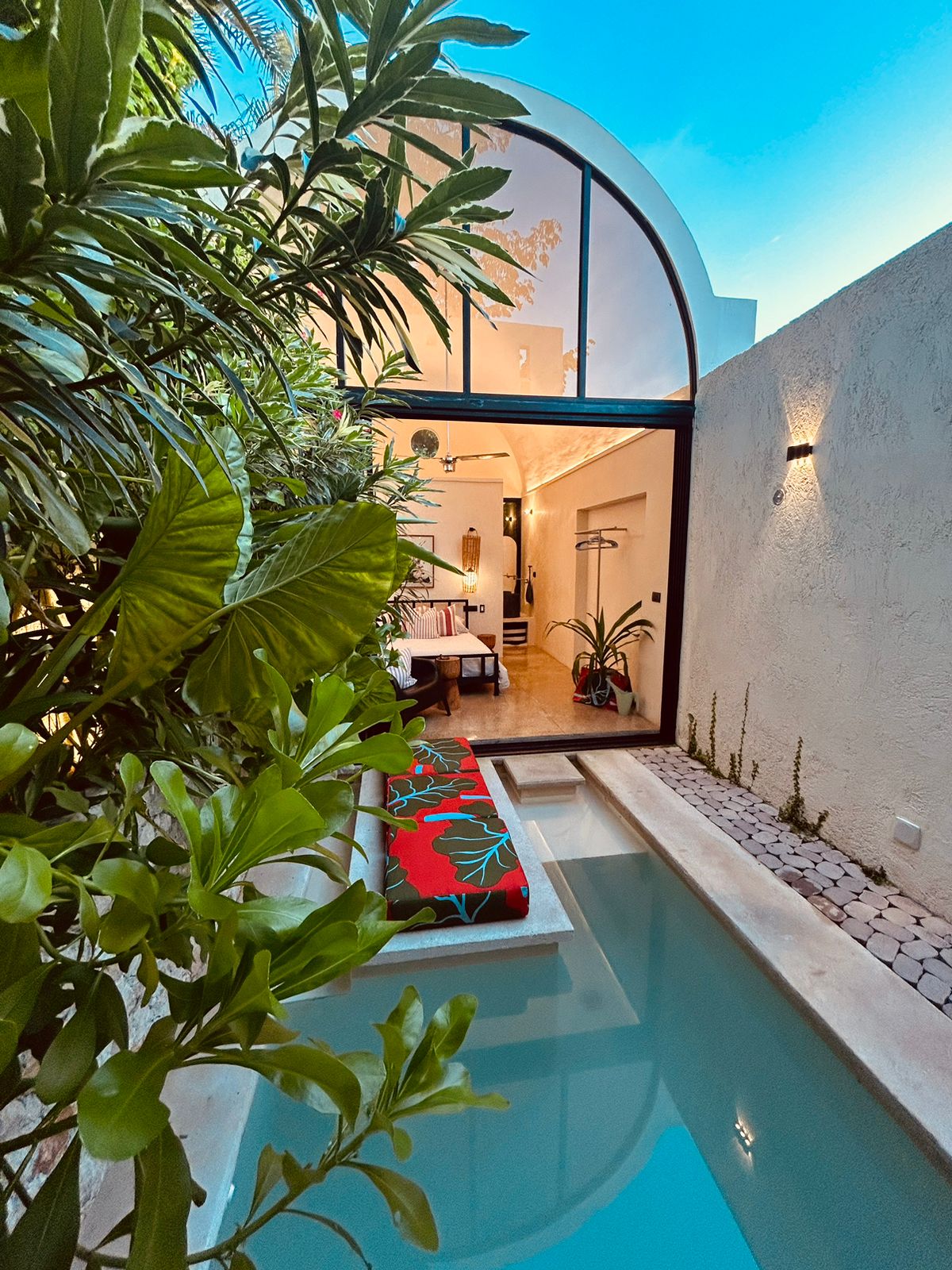 A private pool is framed by tropical plants, offering a glimpse of the inviting interior through large glass doors. The pool reflects the clear sky, while a colorful bench adds a touch of vibrancy to the serene outdoor space.
