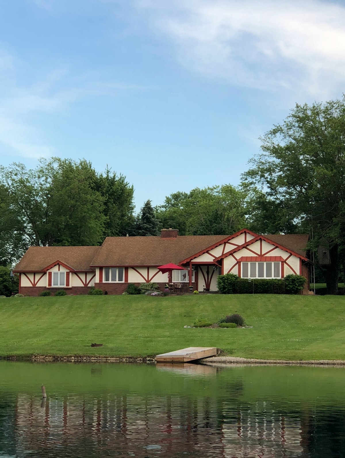 A charming lakefront home is showcased with distinctive architectural details, including a red and white exterior. The property is framed by lush greenery and features multiple windows allowing natural light inside. A spacious lawn leads down to the water's edge, enhancing the serene setting.