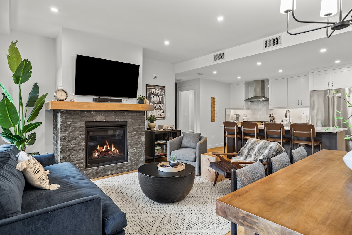 A modern living space features a cozy gas fireplace with a sleek stone facade. Comfortable seating is provided by a plush sofa and armchairs, while a large dining table invites shared meals. Natural light illuminates the area, complemented by contemporary decor and well-defined kitchen in the background.