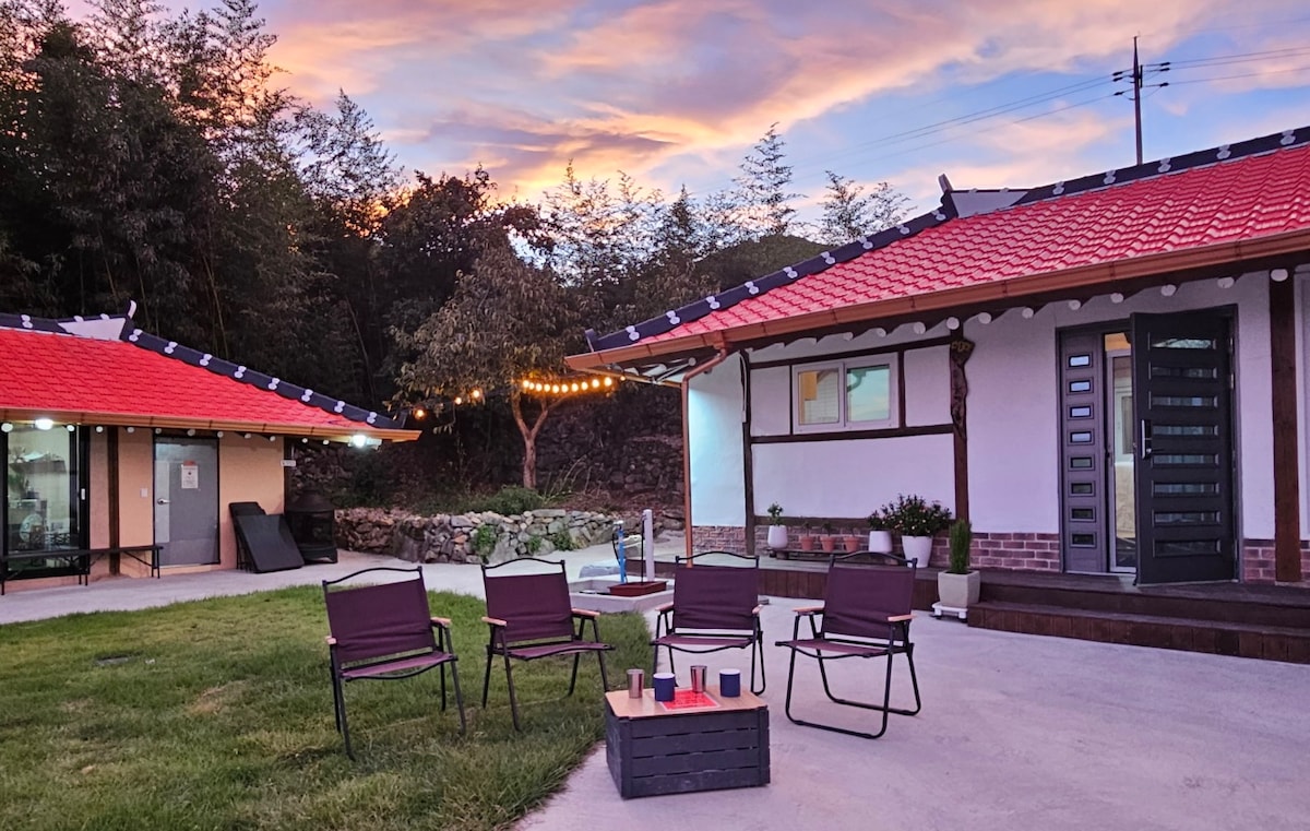 An outdoor space features four folding chairs arranged around a small table. The relaxing area is set against a backdrop of two traditional 한옥 buildings with bright red roofs. Soft lighting enhances the ambiance as a colorful sunset fills the sky.