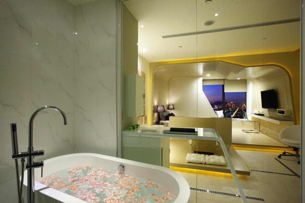A modern bathroom presents a freestanding bathtub filled with water and floating petals. The glass panel offers a view into a contemporary living space featuring soft lighting and a large screen TV, with a cozy seating area visible in the background.