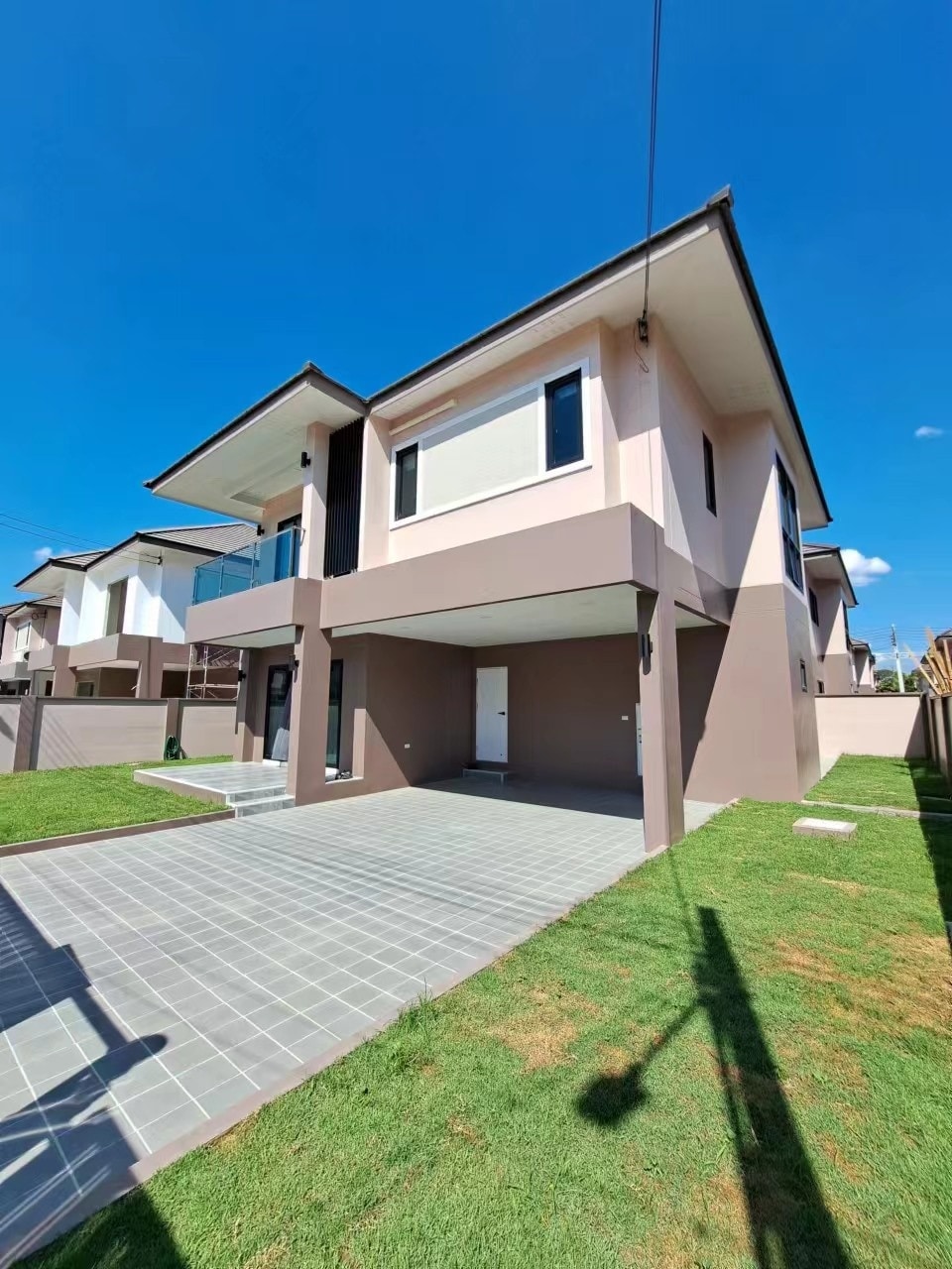 The exterior of the house features a contemporary design with a two-story structure. A spacious driveway and gray tiled pathway lead to the main entrance. The surrounding area is covered in lush green grass under a clear blue sky.