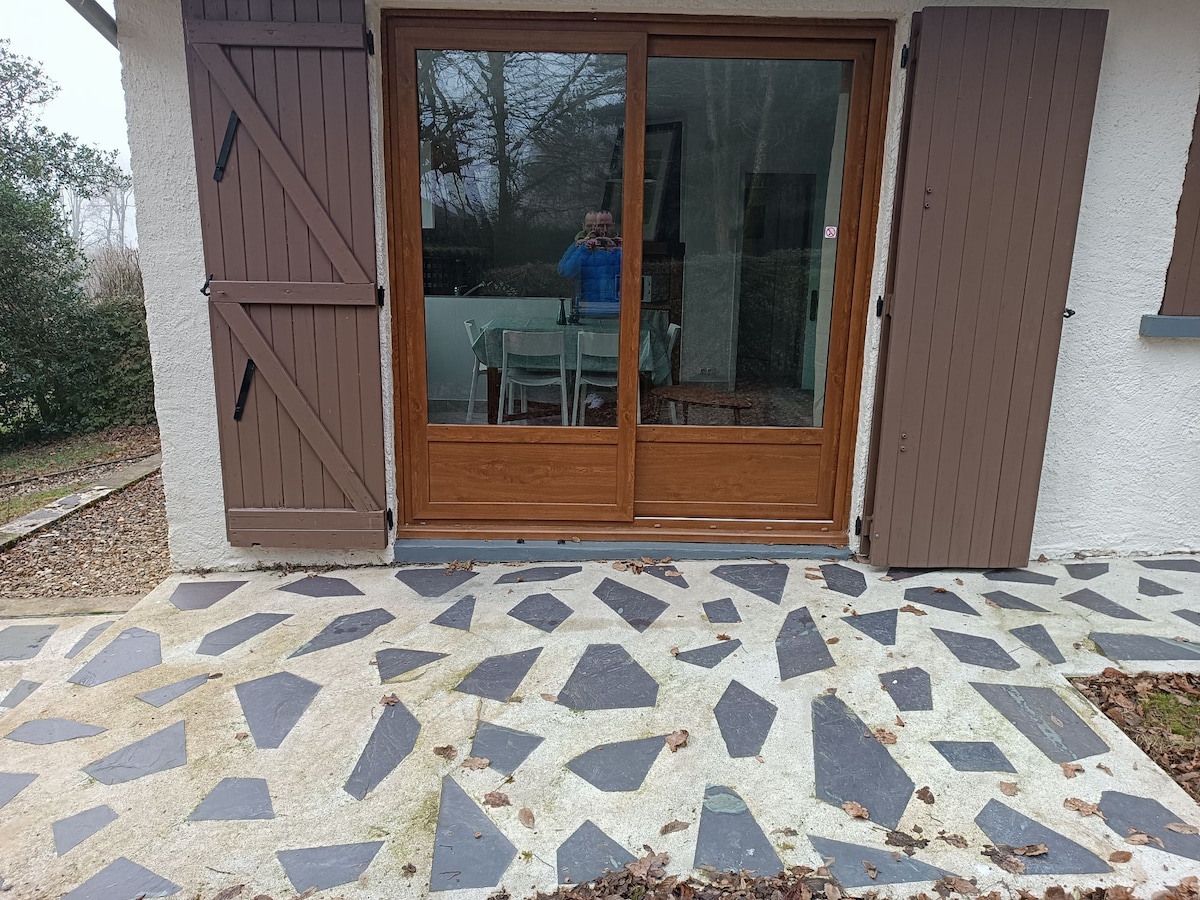 An outdoor patio features a unique stone pattern, along with wooden shutters framing a large glass door. A glimpse of a light-colored dining table can be seen through the glass, extending the inviting space into the interior.