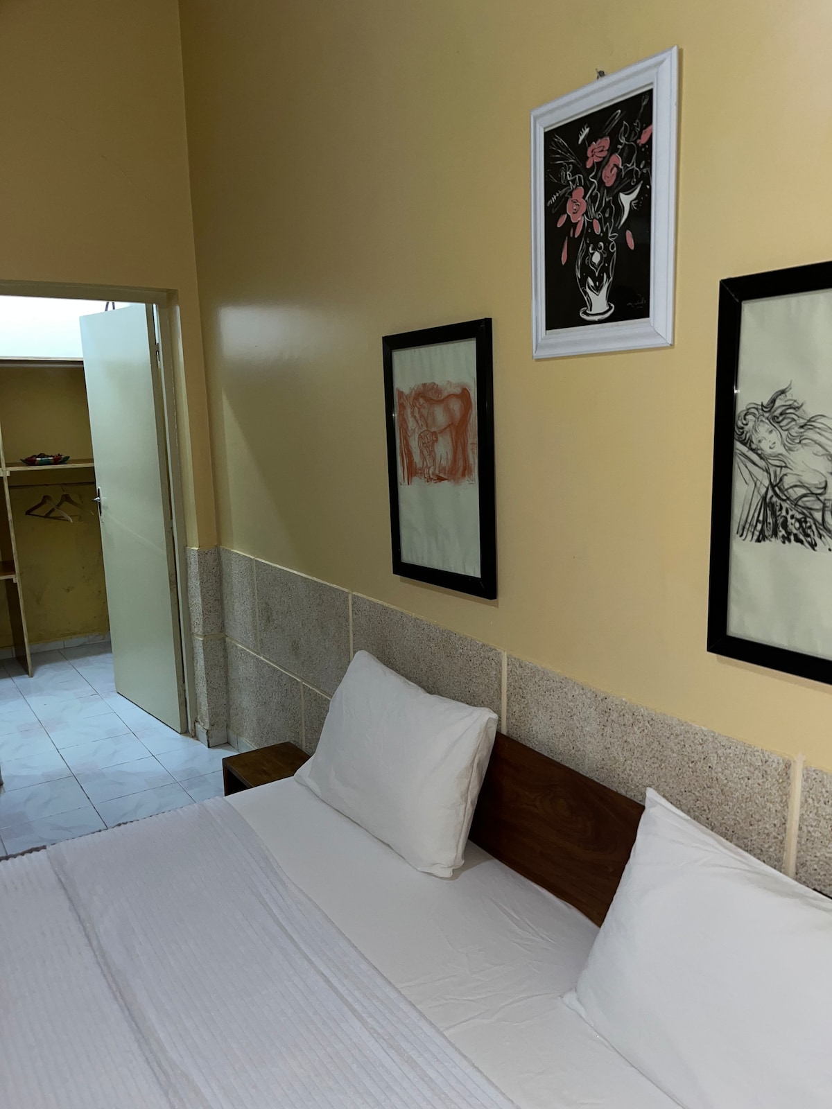 A minimalist bedroom features a spacious bed with white linens, flanked by two framed artworks on the yellow wall. A light-colored wooden headboard adds warmth. A doorway leads to a closet space, while natural light softly illuminates the room from an unseen source.