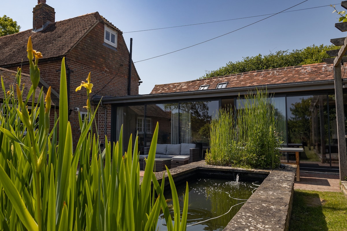 The exterior shows a sleek glass extension of the cowshed, surrounded by tall green plants. A koi carp pond with a gentle fountain sits in the foreground, while a comfortable seating area is visible on the patio adjacent to the main building.