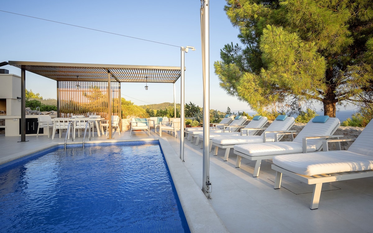 A private swimming pool is surrounded by sun loungers, offering a clear view of the serene landscape. An outdoor dining area under a shaded pergola provides space for gatherings. Natural greenery frames the scene, enhancing the tranquil atmosphere.