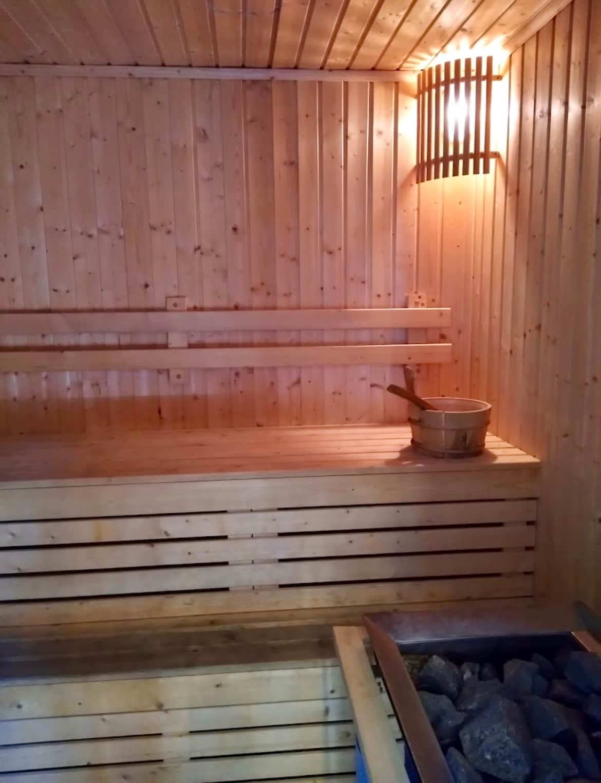 A warm wooden sauna interior is viewed, featuring bench seating along two walls and ambient lighting illuminating the space. A wooden bucket is positioned on one bench, with a stone heater visible, creating a serene atmosphere for relaxation.