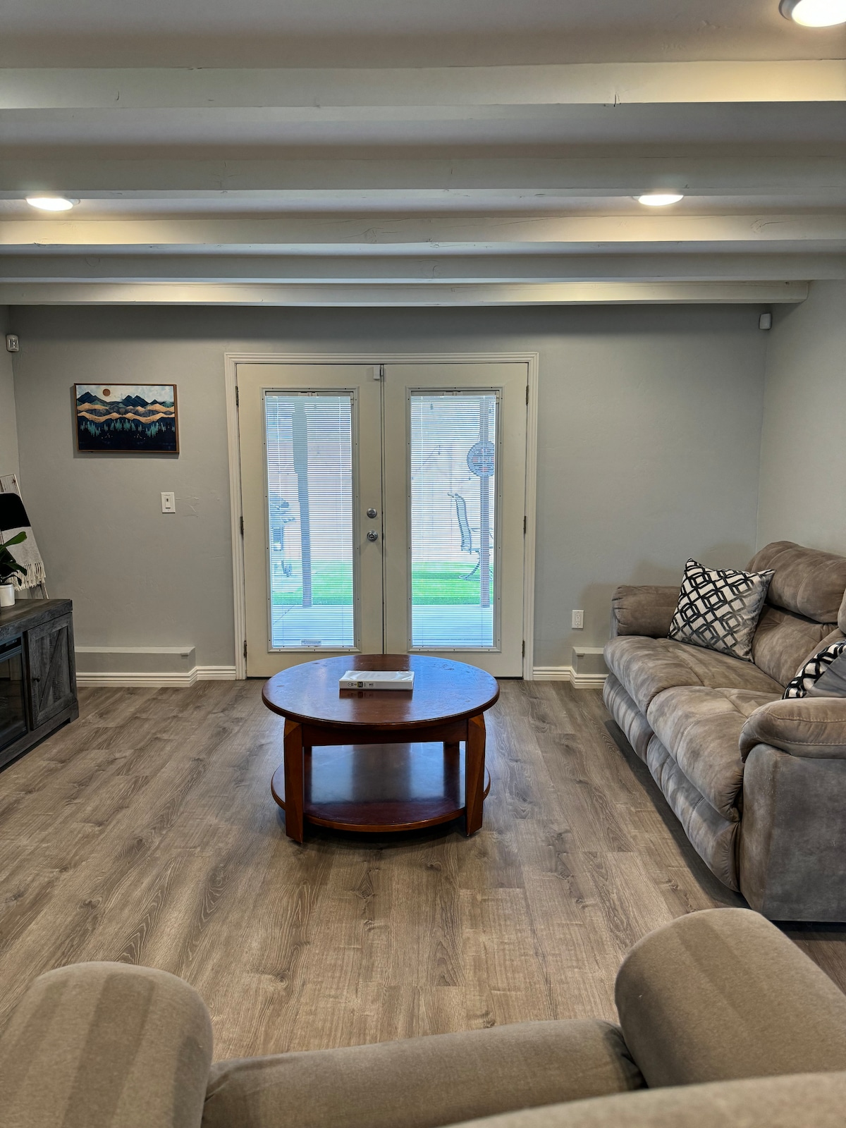 A living room space features comfortable gray sofas complemented by a round wooden coffee table. Large glass doors provide access to an outdoor area, allowing natural light to fill the room. Recessed lighting and wooden beams enhance the ceiling's aesthetic.