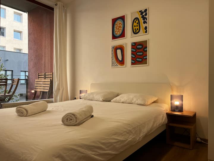 舒适的卧室，配有标准双人床、干净的床单和卷起的毛巾。 充满活力的艺术和柔和的床头照明营造出温馨的氛围，滑动门通往私人阳台。