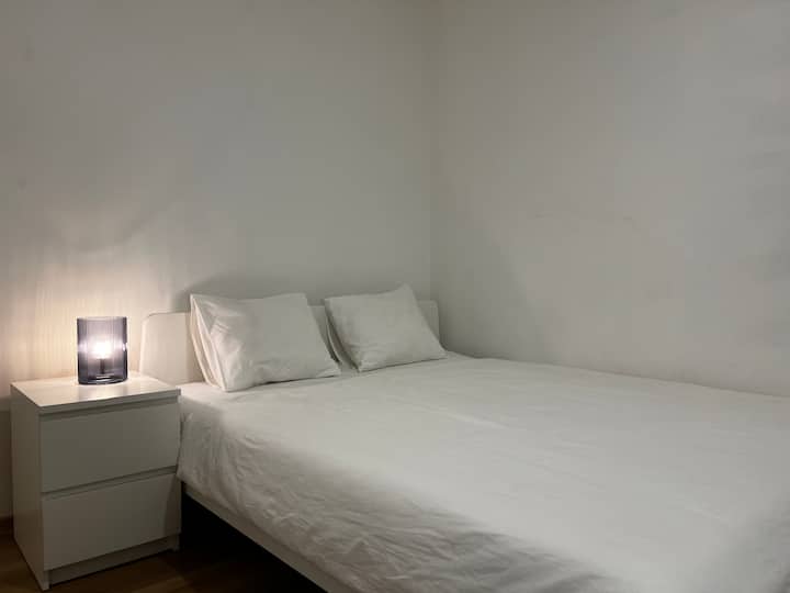 宁静、简约的卧室，配有双人床，铺有洁白的床单和蓬松的枕头；床头柜旁边的柔和灯光营造出温馨的氛围。