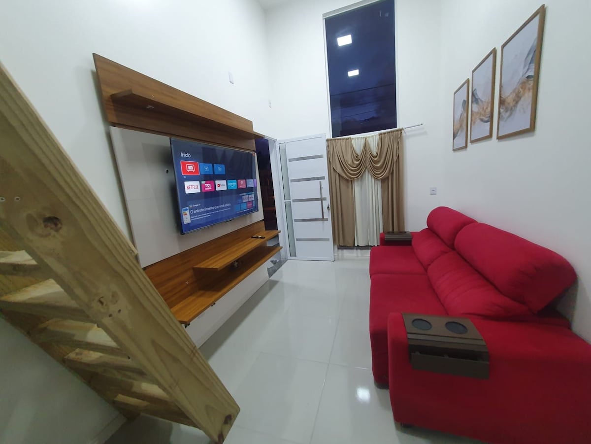 A modern living space is presented, featuring a plush red sofa with integrated cup holders. A large wall-mounted television is centered on a wooden shelf, complemented by decorative artwork above. The room is brightened by natural light filtering through large windows and elegant curtains.