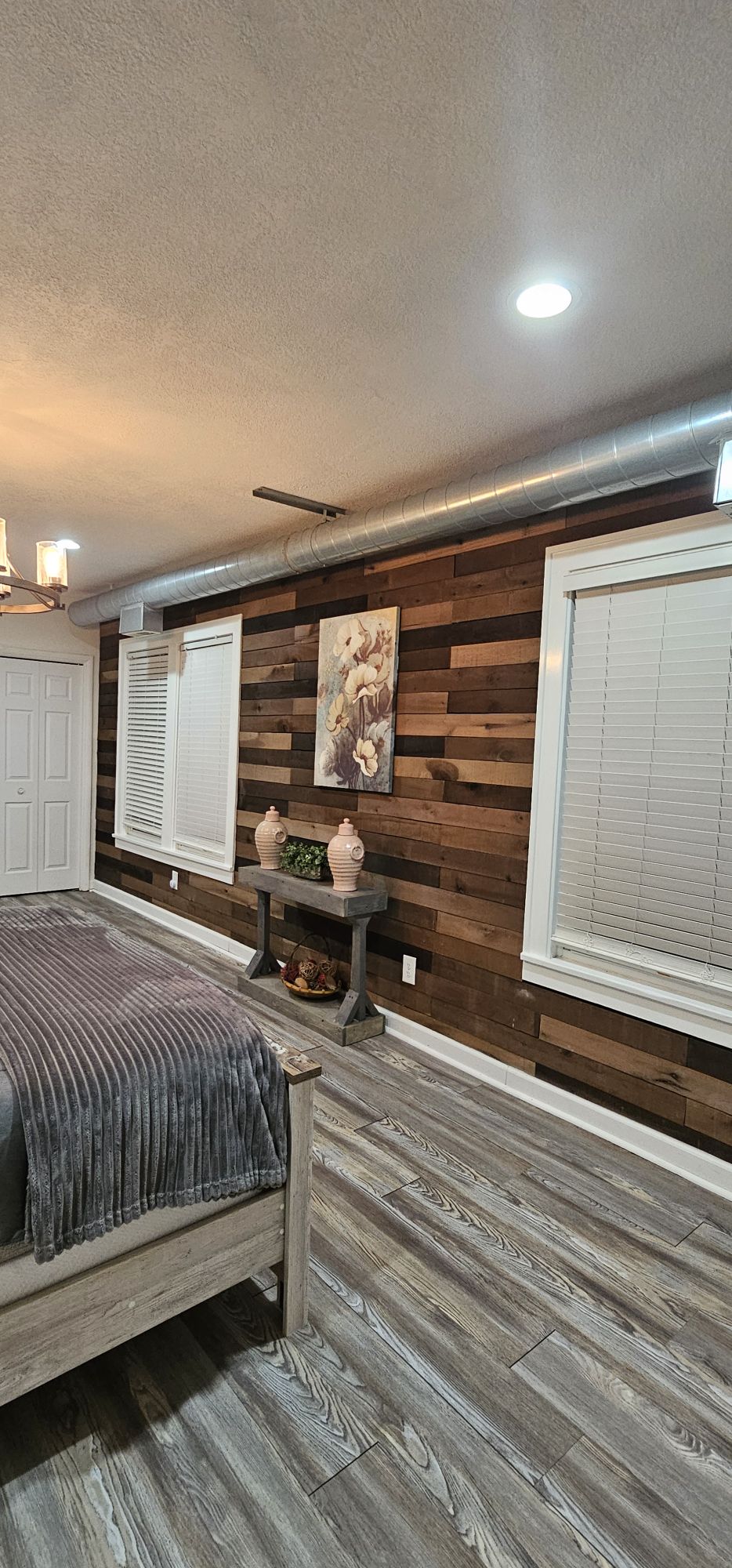 A cozy bedroom is showcased, featuring a wooden accent wall adorned with a floral artwork. A spacious bed is complemented by a textured blanket. Natural light enters through two partially open windows, while a wooden table with decorative vases adds a touch of elegance to the space.