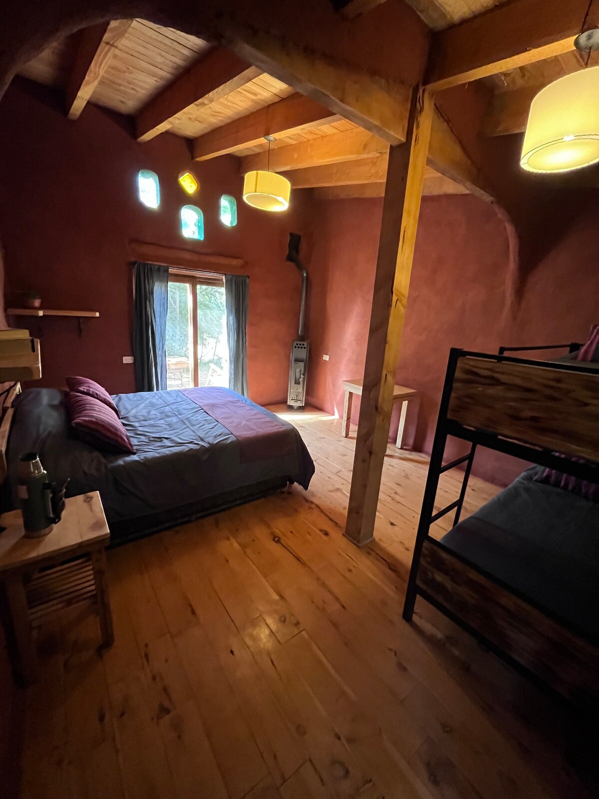 A warm and earthy-toned bedroom features a spacious bed with layered bedding. A cozy bunk bed occupies one corner, complemented by rustic wooden furniture. Natural light enters through a large window, while unique stained glass accents add color to the walls.