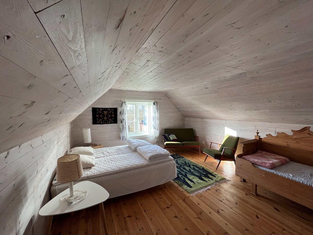 An airy attic room features sloped wooden ceilings and light-toned walls. A spacious bed is positioned alongside a single bed, with a small table and lamp nearby. Natural light enters through a window adorned with light curtains, creating a bright and comfortable space.