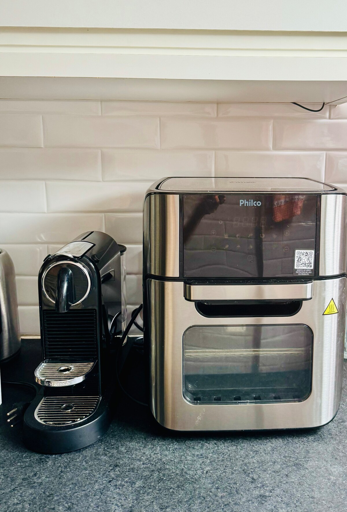 A modern kitchen countertop is shown, featuring a sleek Philco coffee maker alongside a compact espresso machine. The appliances are positioned against a backdrop of light-colored tiled backsplash, complemented by a dark countertop. The area is designed for convenience and functionality.