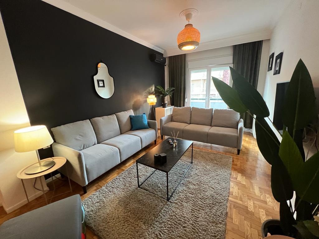 The living room features two soft grey sofas positioned around a low coffee table on a textured rug. Natural light enters through a glass door leading to an outdoor space, with indoor plants adding a touch of greenery to the warm atmosphere.