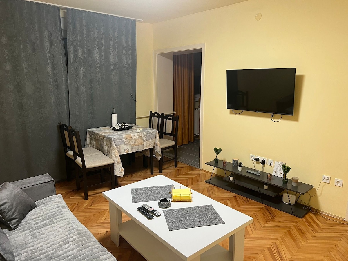 A living area features a gray sofa adjacent to a white coffee table. A dining table with chairs is placed in the corner, and a wall-mounted flat-screen television is present. Soft curtains frame the windows, adding a touch of warmth to the neutral-colored walls.