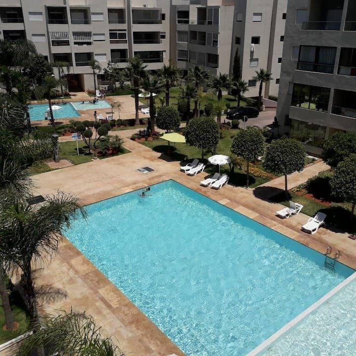 The image showcases a serene pool area surrounded by well-maintained gardens. Two large swimming pools are visible, accompanied by lounge chairs and palm trees, providing a tranquil setting for relaxation. Apartment buildings can be seen in the background, creating a peaceful residential atmosphere.