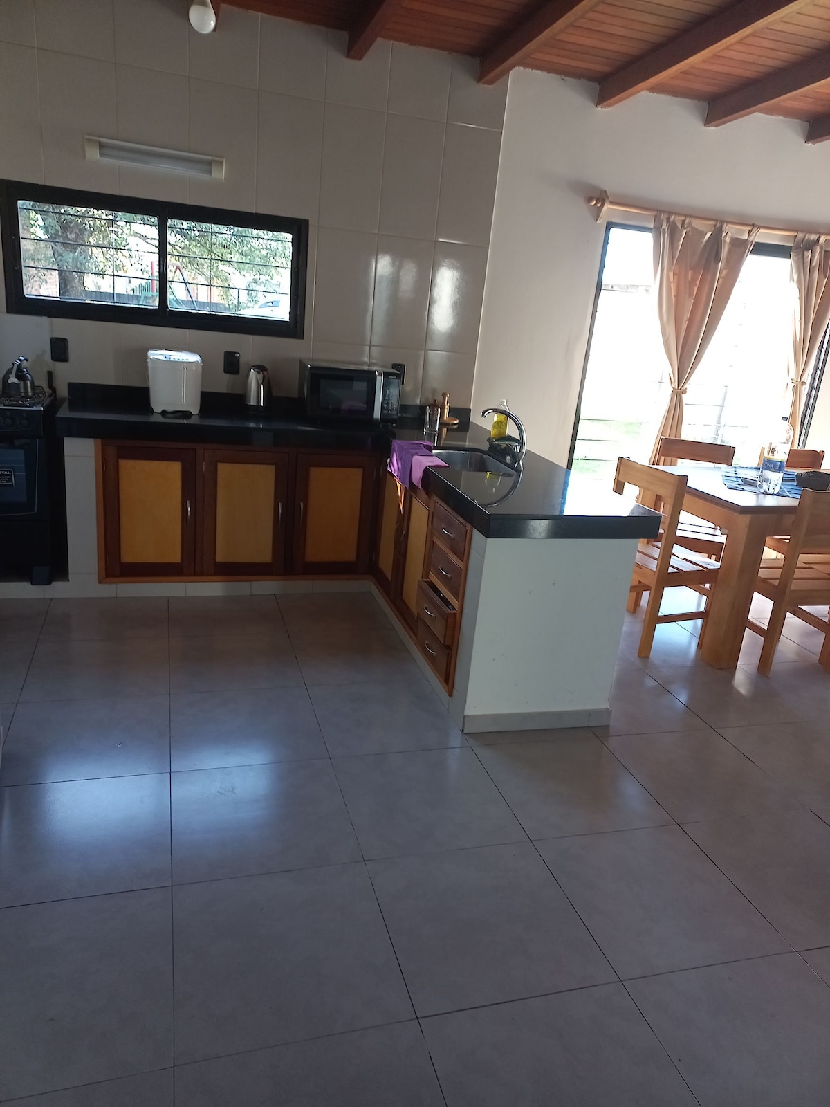The kitchen area features wooden cabinetry and a black countertop. Essential appliances, including a microwave and a kettle, are visible. A dining table with wooden chairs is positioned near large windows that allow natural light to illuminate the space.