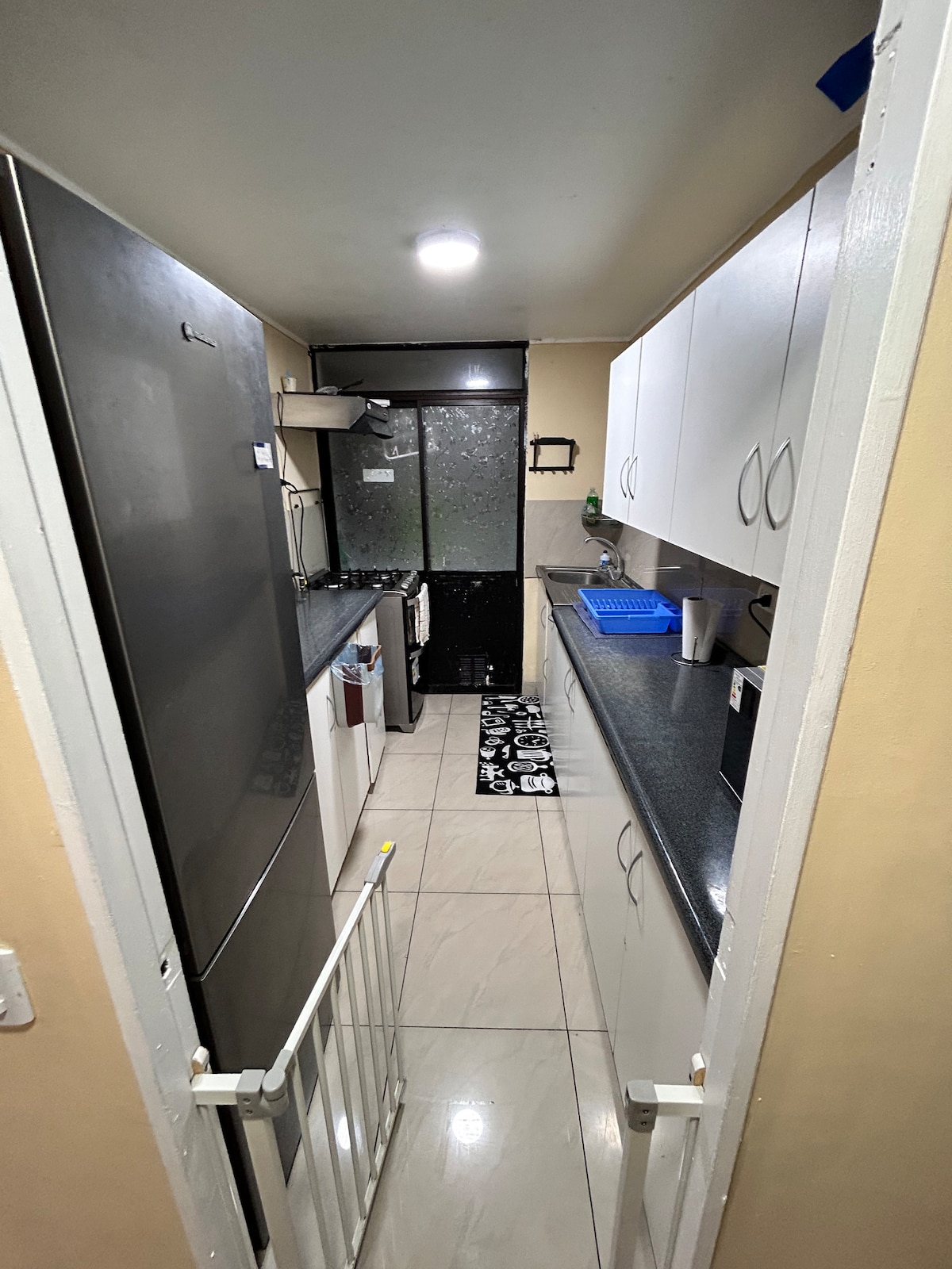 A narrow kitchen with sleek black countertops and white cabinetry is presented. Stainless steel appliances are positioned along one side, while a small portable gate is placed at the entrance. A modern rug is located on the tiled floor, adding a touch of comfort.