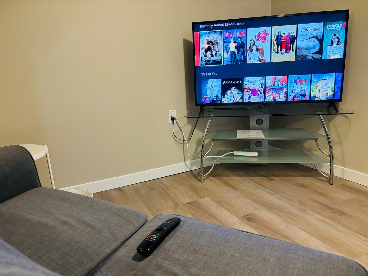 A spacious living area features a large flat-screen TV displaying a selection of movies, complemented by a sleek glass TV stand. A remote control sits on a soft, neutral-colored sofa, enhancing the comfortable environment. Warm, natural light filters softly into the space.