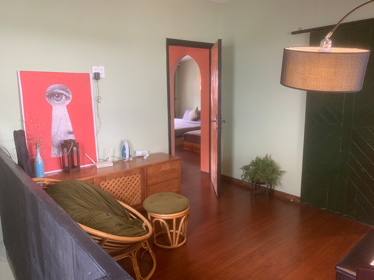 A welcoming living space features a combination of modern and classic design elements. A vibrant artwork adorns the wall, while a comfortable chair and small table are positioned beside a dark green door. Wooden flooring enhances the overall warmth of the room.
