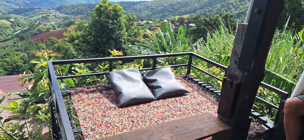 A cozy outdoor seating area features a wooden platform adorned with colorful textiles and two plush pillows. The surrounding greenery and distant hills create a serene view, providing a perfect spot for relaxation and enjoying nature.