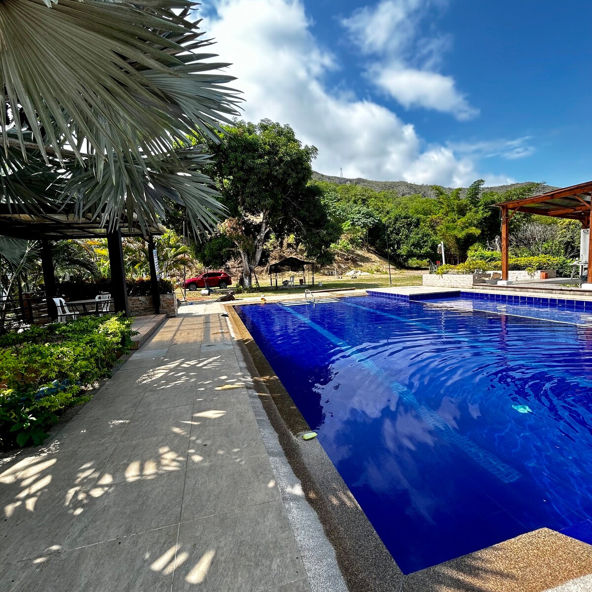 A clear blue pool reflects the sky, surrounded by a spacious patio and tropical landscaping. Nearby, a shaded seating area offers a place to relax, complemented by lush greenery and distant hills. Sunlight creates gentle patterns on the pool's surface.