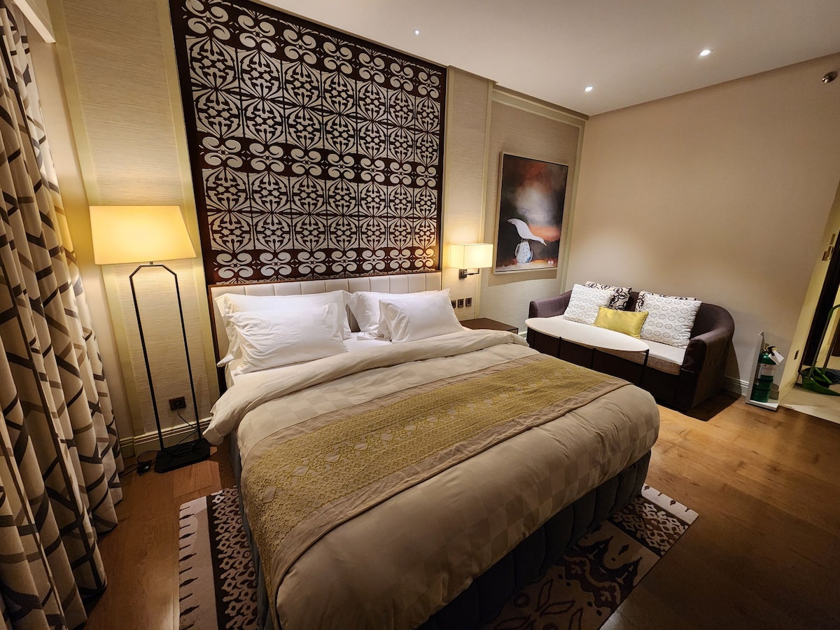 A stylish bedroom features a spacious bed with crisp white linens and a textured yellow throw. A patterned wall serves as a focal point, while a cozy seating area is positioned beside the bed. Soft lighting and wooden flooring create a calming environment.