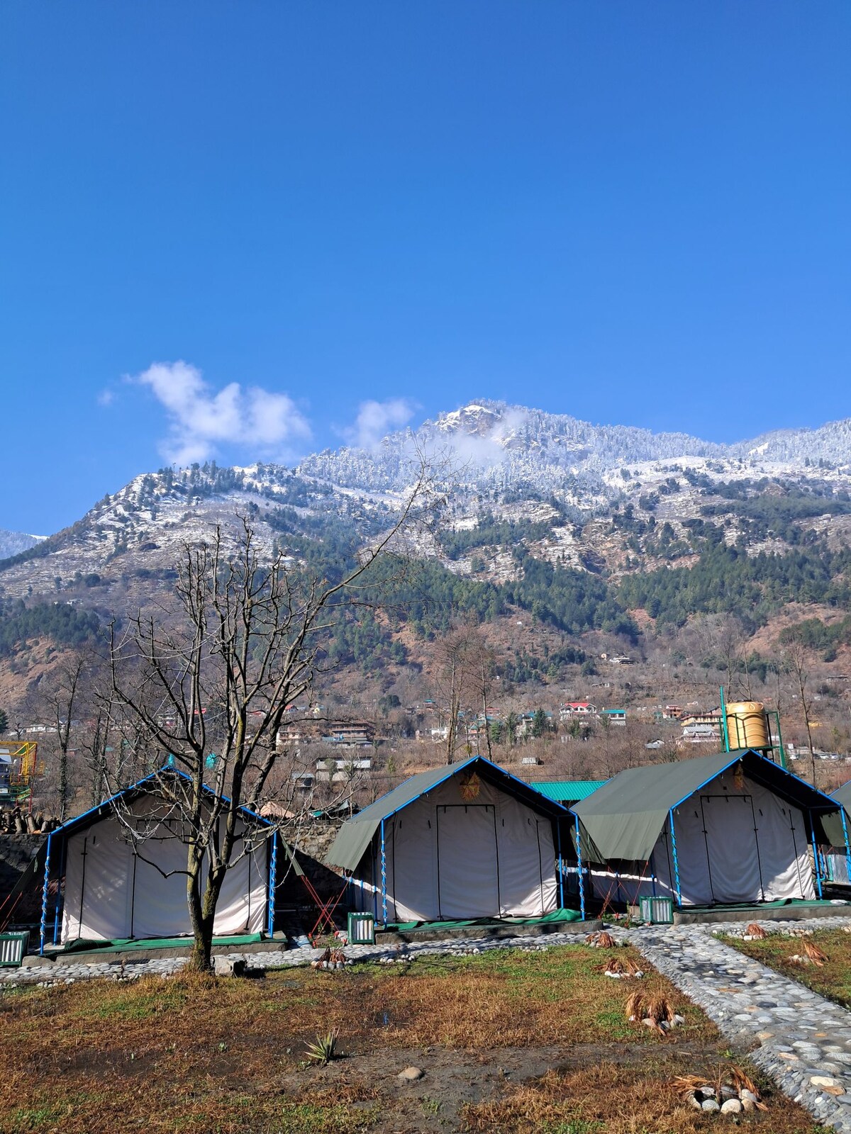 A cluster of canvas tents is set against a backdrop of snow-capped mountains. The area is surrounded by grass and a few trees, with a clear blue sky overhead. A stone path leads to the tents, enhancing the serene outdoor setting.