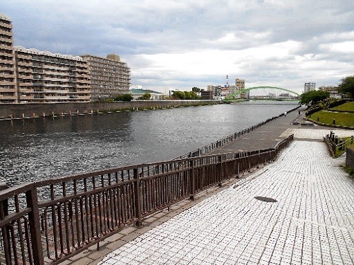 A river scene is depicted, featuring calm waters bordered by a walkway. On one side, a metal railing runs alongside the path, while residential buildings rise in the background. A bridge can be seen in the distance, adding a sense of connection to the landscape.