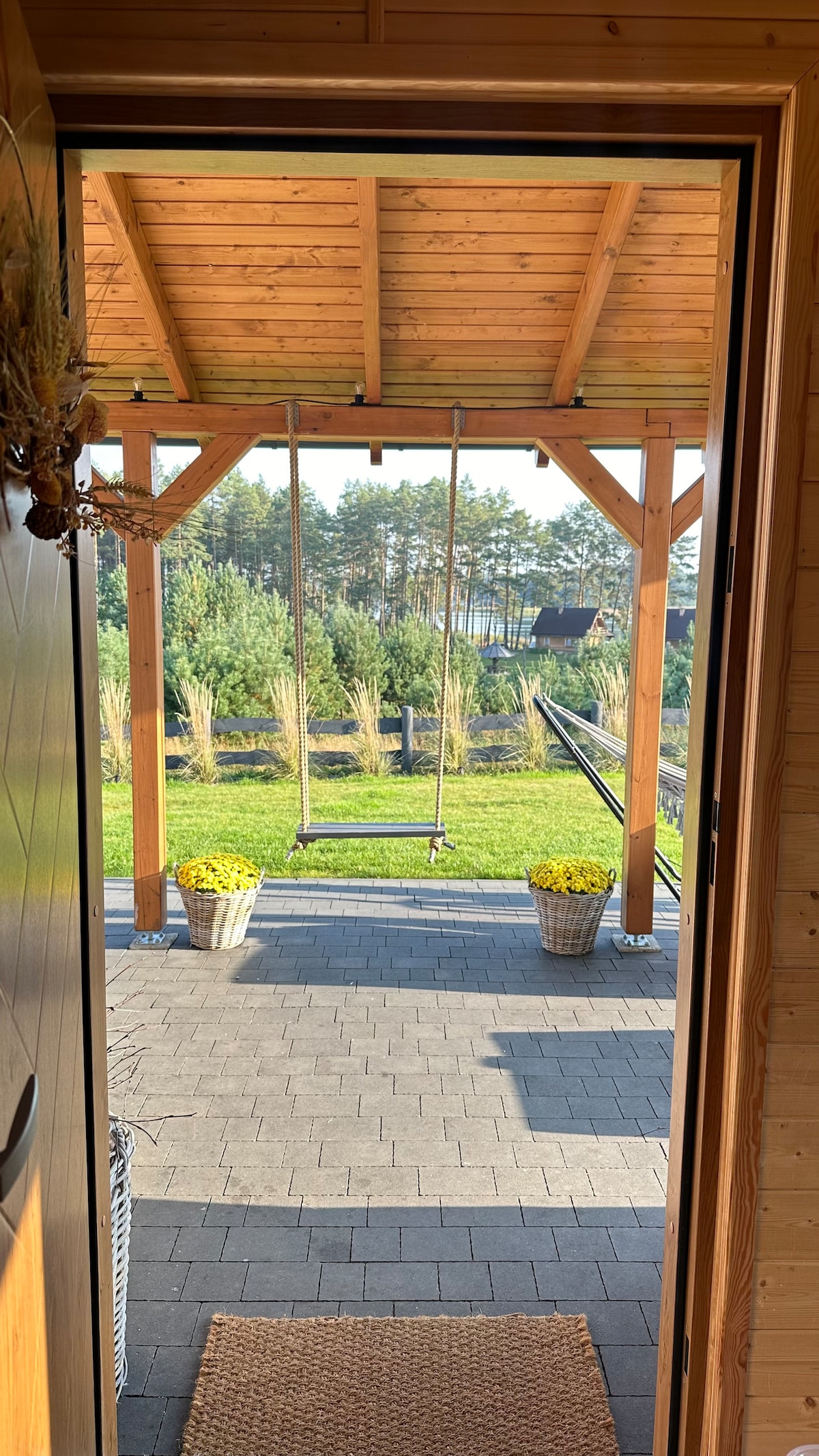 A wooden door stands open, revealing a porch with a swing. Potted yellow flowers sit on either side, while a neatly paved pathway leads to a grassy area beyond. Trees and a glimpse of the lake are visible in the background.