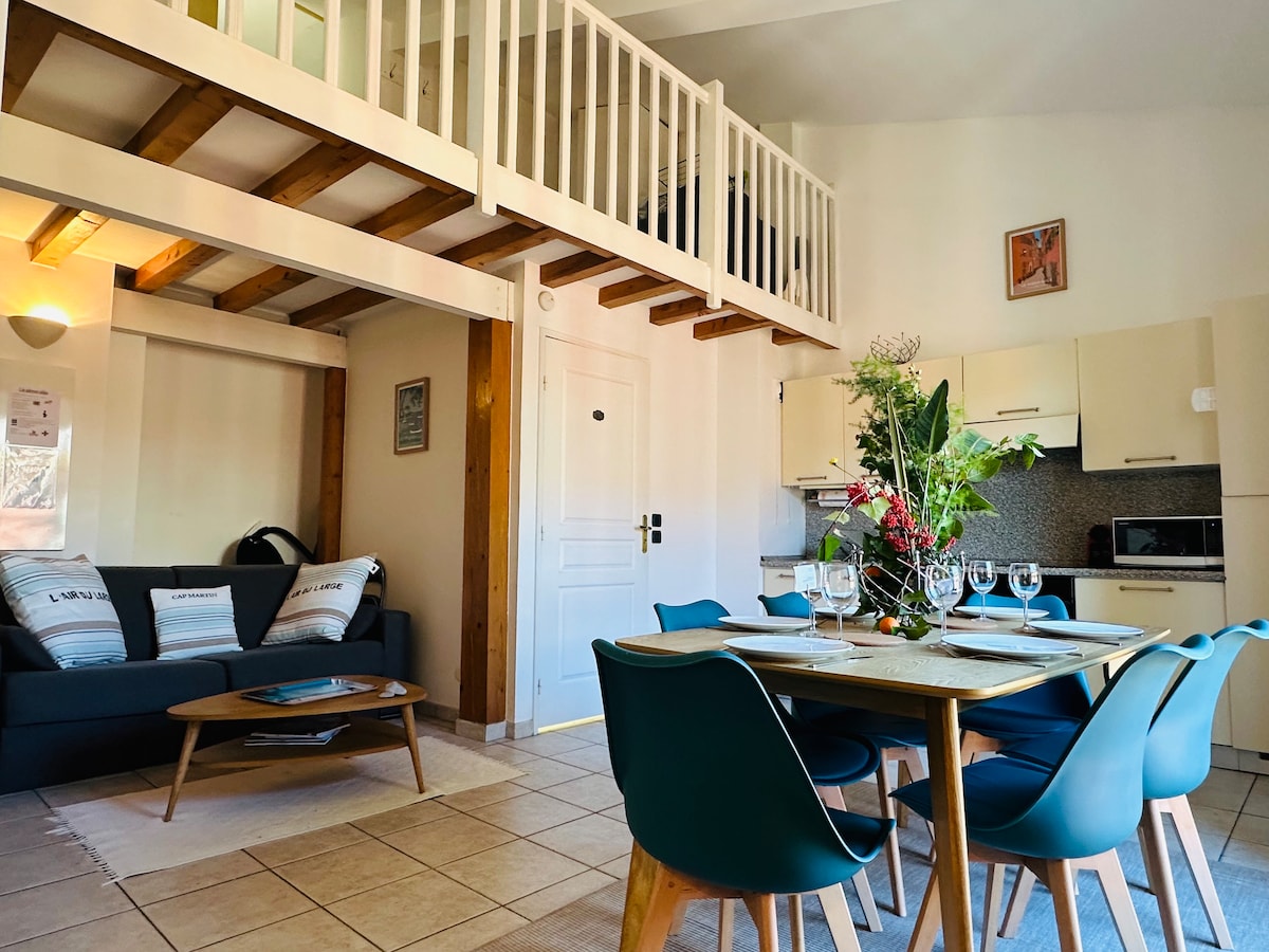 A cozy living area features a dark sofa and a wooden coffee table, complemented by a dining table surrounded by blue chairs. The kitchen area is equipped with modern appliances. A loft with a safety railing is visible above, creating an open and inviting layout.