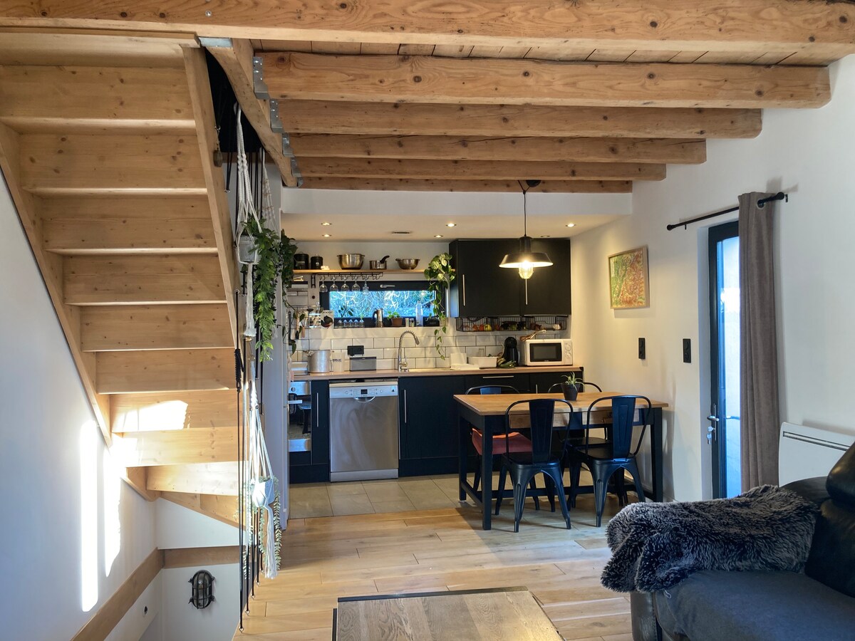 An open-concept living area is highlighted by exposed wooden beams. A dining table with black chairs sits adjacent to a modern kitchen, equipped with sleek appliances. Natural light filters in through a glass door leading outdoors, enhancing the spaciousness of the room.